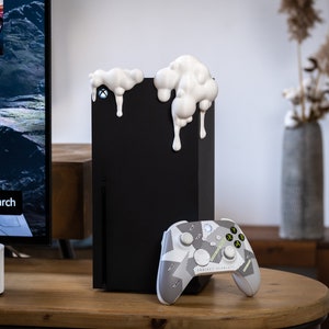 Może przedstawiać: Czarna konsola Xbox Series X z białymi akcentami z przodu. Konsola stoi na drewnianej powierzchni obok biało-szarego kontrolera Xbox w kamuflażu.