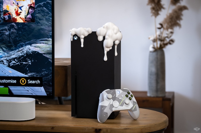 Może przedstawiać: Czarna konsola Xbox Series X z białymi akcentami z przodu. Konsola stoi na drewnianej powierzchni obok biało-szarego kontrolera Xbox w kamuflażu.