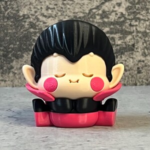 K&ouml;nnte beinhalten: Eine 3D-gedruckte Figur eines Cartoon-Vampirs mit geschlossenen Augen und rosigen Wangen. Die Figur hat schwarzes Haar, einen schwarzen Kragen und einen rosa Umhang und K&ouml;rper. Die Figur sitzt mit verschr&auml;nkten H&auml;nden.