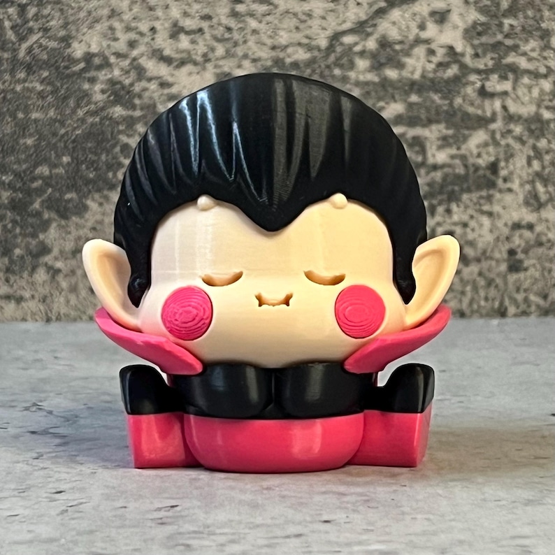 K&ouml;nnte beinhalten: Eine 3D-gedruckte Figur eines Cartoon-Vampirs mit geschlossenen Augen und rosigen Wangen. Die Figur hat schwarzes Haar, einen schwarzen Kragen und einen rosa Umhang und K&ouml;rper. Die Figur sitzt mit verschr&auml;nkten H&auml;nden.