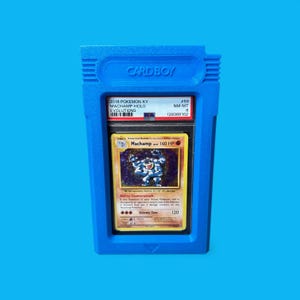 May include: A blue "CARD BOY" case holding a graded Pok&eacute;mon card. The card features a Machamp illustration and text. The case has the text "CARD BOY" at the top and is set against a blue background.