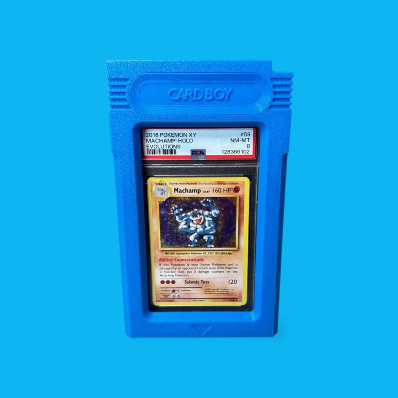 May include: A blue "CARD BOY" case holding a graded Pok&eacute;mon card. The card features a Machamp illustration and text. The case has the text "CARD BOY" at the top and is set against a blue background.
