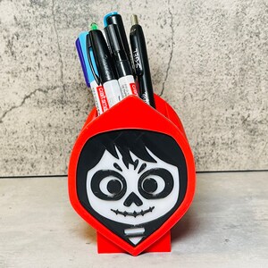 May include: Red plastic pencil holder shaped like the head of a character from the movie Coco. The character has black hair, a white face, and a black skull design. The holder has four slots for pens or pencils.