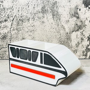 May include: White desk organizer shaped like a monorail train with black and red details. The organizer has three compartments.