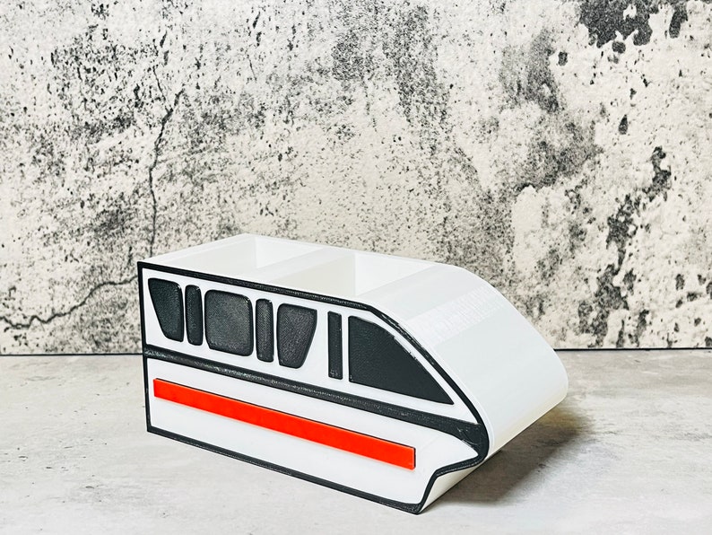 May include: White desk organizer shaped like a monorail train with black and red details. The organizer has three compartments.