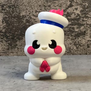 K&ouml;nnte beinhalten: Eine wei&szlig;e Figur eines Zeichentrickcharakters mit Matrosenm&uuml;tze und rosa Akzenten auf den Wangen und der Fliege. Der Hut hat ein blaues Band und eine rosa Krone. Die Figur hat schwarze Augen und ein kleines L&auml;cheln. Die Figur steht auf einer grauen Oberfl&auml;che.