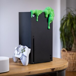 Może przedstawiać: Czarna konsola Xbox Series X z zielonym, śluzowatym obiektem na g&oacute;rze. Biały i szary kontroler Xbox leży na stole obok konsoli.
