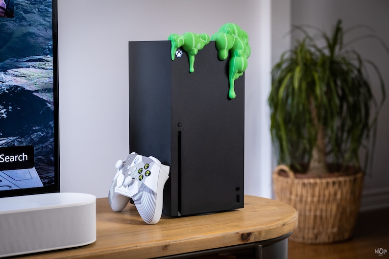 Może przedstawiać: Czarna konsola Xbox Series X z zielonym, śluzowatym obiektem na g&oacute;rze. Biały i szary kontroler Xbox leży na stole obok konsoli.