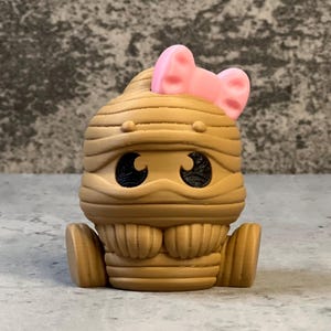 K&ouml;nnte beinhalten: Eine hellbraune Mumienfigur mit einer rosa Schleife auf dem Kopf. Die Mumie hat gro&szlig;e schwarze Augen und sitzt mit angewinkelten Beinen. Die Figur besteht aus einem festen Material und hat eine strukturierte Oberfl&auml;che.