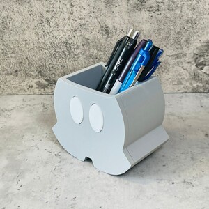 K&ouml;nnte beinhalten: Ein grauer, 3D-gedruckter Stiftehalter in Form eines Gespenstes mit zwei wei&szlig;en Augen. Der Halter ist mit Stiften und Bleistiften gef&uuml;llt.