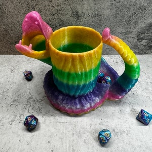May include: A rainbow-coloured, 3D printed dragon mug with a handle shaped like a tail. The mug is sitting on a table with several blue and purple dice.