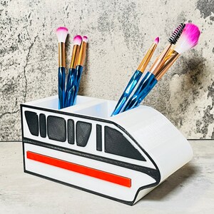 May include: White ceramic makeup brush holder shaped like a monorail train with a red stripe. The holder has two compartments for brushes.  The holder has black and gray details.  Five makeup brushes are in the holder, including pink and blue brushes.