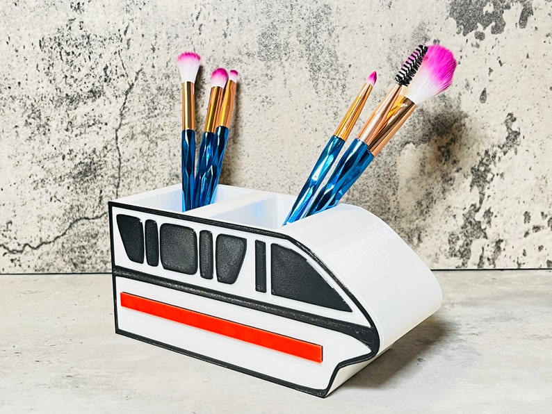 May include: White ceramic makeup brush holder shaped like a monorail train with a red stripe. The holder has two compartments for brushes.  The holder has black and gray details.  Five makeup brushes are in the holder, including pink and blue brushes.