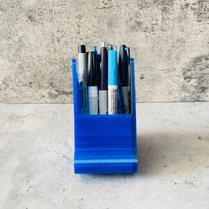May include: A blue plastic pen holder with six pens inside. The pens are black, blue, and white.