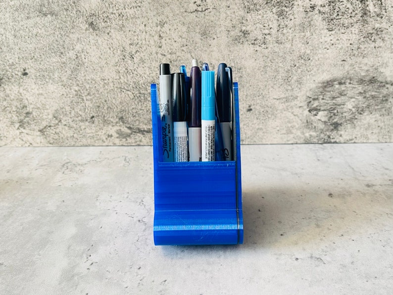 May include: A blue plastic pen holder with six pens inside. The pens are black, blue, and white.