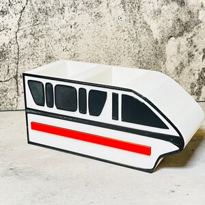 May include: White desk organizer shaped like a monorail train with black windows and a red stripe.