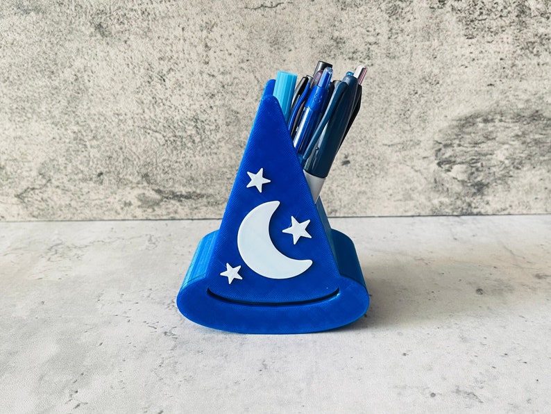 May include: A blue 3D printed pen holder shaped like a wizard's hat with a crescent moon and stars design. The holder is filled with pens and pencils.