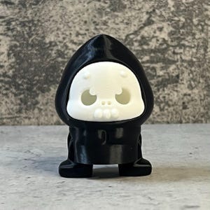 K&ouml;nnte beinhalten: Kleine, schwarze Figur einer Kapuzengestalt mit wei&szlig;em Totenkopfgesicht. Die Figur hat eine abgerundete Kapuze und ein einfaches, cartoonhaftes Design. Das Totenkopfgesicht hat gro&szlig;e Augenh&ouml;hlen und einen kleinen Mund.