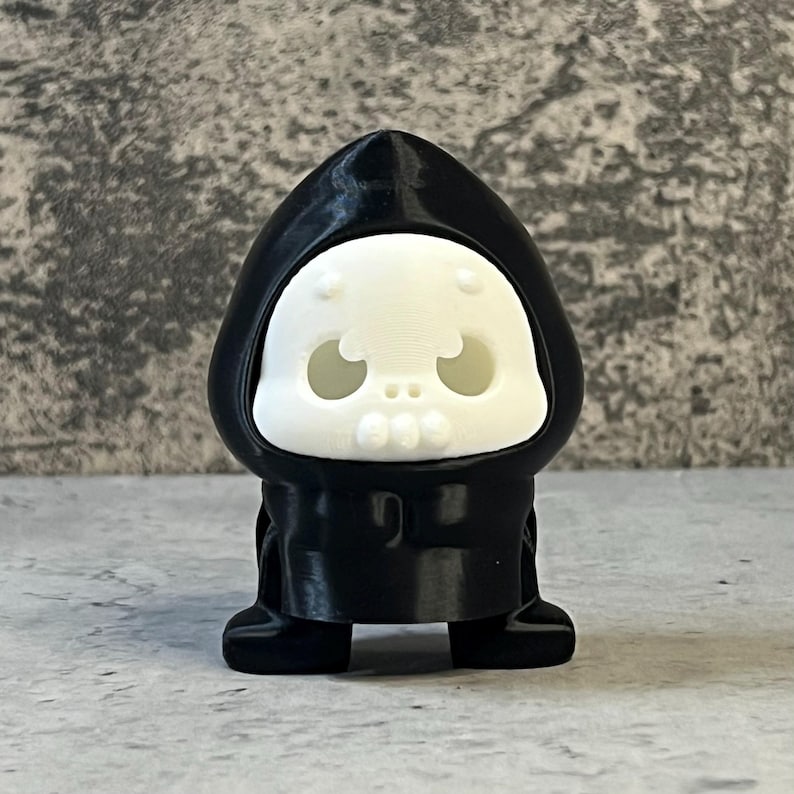 K&ouml;nnte beinhalten: Kleine, schwarze Figur einer Kapuzengestalt mit wei&szlig;em Totenkopfgesicht. Die Figur hat eine abgerundete Kapuze und ein einfaches, cartoonhaftes Design. Das Totenkopfgesicht hat gro&szlig;e Augenh&ouml;hlen und einen kleinen Mund.