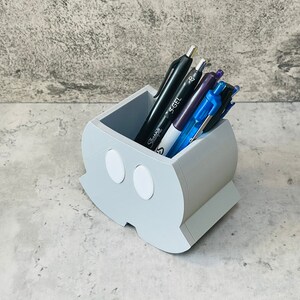 K&ouml;nnte beinhalten: Ein grauer, 3D-gedruckter Stiftehalter in Form eines Gespenstes mit zwei wei&szlig;en Augen. Der Halter ist mit verschiedenen Stiften gef&uuml;llt, darunter schwarze, violette und blaue.
