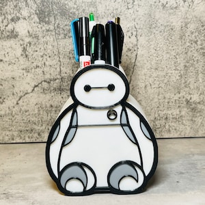 Könnte beinhalten: Weißer Plastik-Stiftbehälter in Form von Baymax aus dem Film Big Hero 6. Der Behälter hat schwarze Umrisse und graue Details. Er enthält mehrere Stifte und Marker.