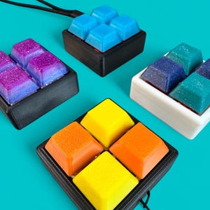 May include: A collection of colorful, square-shaped keycaps in various colors, including blue, purple, yellow, and orange. Each set of keycaps is arranged in a 2x2 configuration and set in a black or white base. The keycaps have a glossy finish.