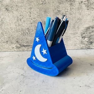May include: A blue wizard hat shaped pencil holder with a white crescent moon and stars design. The holder is filled with pens and pencils.