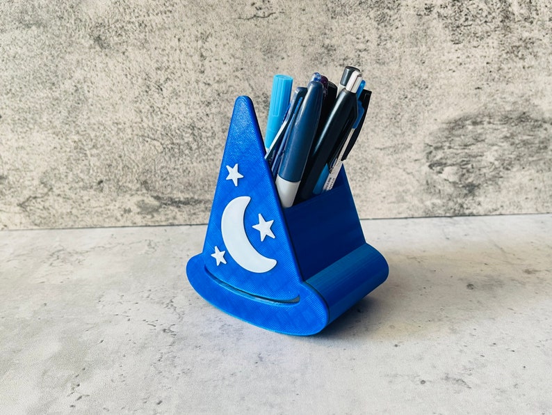 May include: A blue wizard hat shaped pencil holder with a white crescent moon and stars design. The holder is filled with pens and pencils.