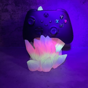 Puede incluir: Un mando de Xbox negro se encuentra sobre una base de cristal brillante multicolor. La base tiene forma de un grupo de cristales y brilla con luz rosa, amarilla y blanca.