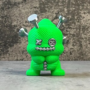 K&ouml;nnte beinhalten: Eine neongr&uuml;ne Figur mit einem gen&auml;hten Design, silbernen N&auml;geln und einem Knopfauge. Die Figur hat einen gen&auml;hten Mund und eine kreuzf&ouml;rmige Naht f&uuml;r ein Auge. Die Figur hat einen gen&auml;hten Torso und Beine.