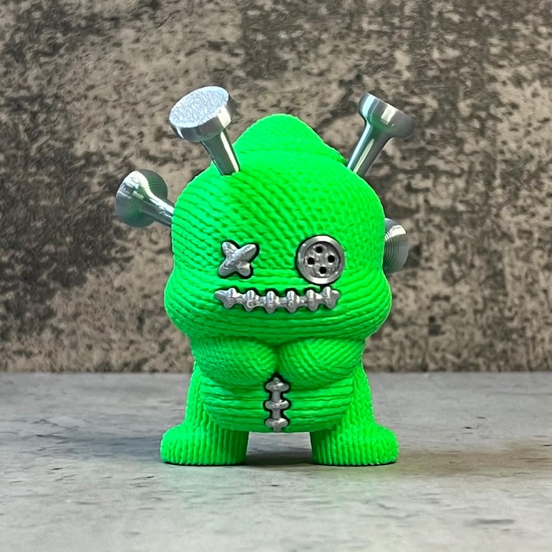 K&ouml;nnte beinhalten: Eine neongr&uuml;ne Figur mit einem gen&auml;hten Design, silbernen N&auml;geln und einem Knopfauge. Die Figur hat einen gen&auml;hten Mund und eine kreuzf&ouml;rmige Naht f&uuml;r ein Auge. Die Figur hat einen gen&auml;hten Torso und Beine.