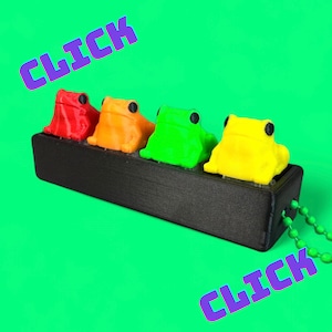 Könnte beinhalten: Ein schwarzes rechteckiges Objekt mit vier bunten Froschfiguren in Rot, Orange, Grün und Gelb. Die Frösche haben schwarze Augen und sind an einer schwarzen Basis befestigt. Das Wort "CLICK" ist in Lila diagonal über das Bild geschrieben.