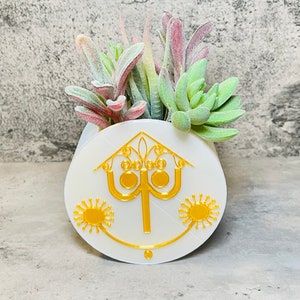 May include: White ceramic planter with a gold design of a stylized house with a lamppost and two suns. The planter is filled with green and red succulents.
