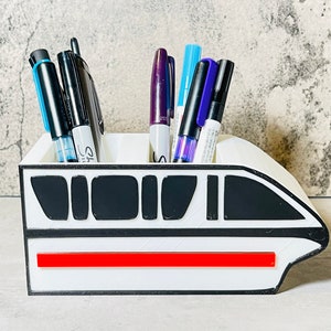 May include: White desk organizer shaped like a monorail with a red stripe. The organizer has black accents and is filled with pens and markers.