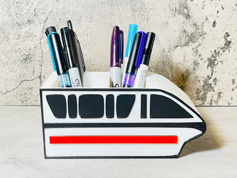 May include: White desk organizer shaped like a monorail with a red stripe. The organizer has black accents and is filled with pens and markers.