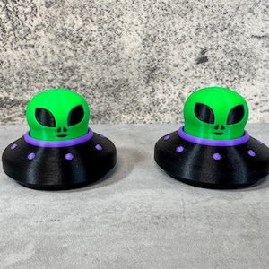 May include: Two green alien figurines with black eyes and faces, atop black saucers with purple accents and purple dots. The alien heads are bright green, and the saucers are black.
