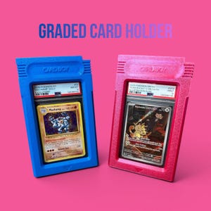 May include: Two graded card holders, one blue and one pink, each holding a Pok&eacute;mon trading card. The blue holder features a Machamp card, while the pink holder displays a Team Rocket's Meowth card. The text "GRADED CARD HOLDER" is at the top.