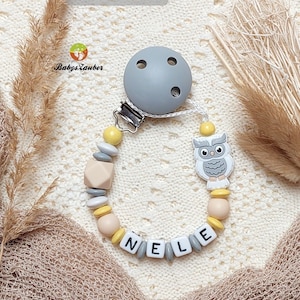 Könnte beinhalten: Ein grauer und gelber Beißring für Babys mit einem Silikon-Schnuller, einem Holz-Eulen-Anhänger und Holzperlen, die den Namen "Nele" buchstabieren.