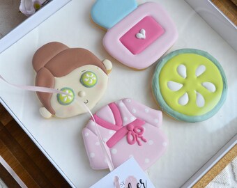 May include: A spa-themed gift box with four decorated cookies. The cookies include a face with cucumber slices on the eyes, a pink robe, a cucumber slice, and a pink and blue perfume bottle. A card reads "Best Mother in the World".