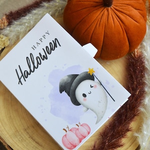 May include: A rectangular white greeting card with the text Happy Halloween in black script. The card features a cartoon ghost wearing a black witch hat and holding a yellow star wand, with two small pink pumpkins at the bottom and a purple watercolor splash.