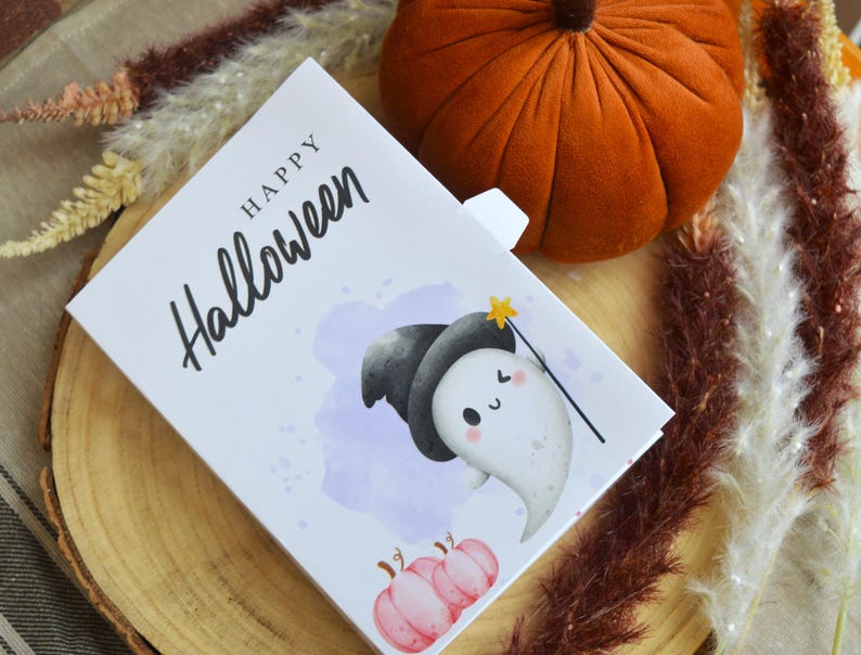 May include: A rectangular white greeting card with the text Happy Halloween in black script. The card features a cartoon ghost wearing a black witch hat and holding a yellow star wand, with two small pink pumpkins at the bottom and a purple watercolor splash.