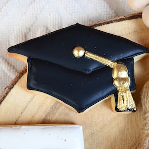 Pu&ograve; includere: Biscotto a forma di cappello di laurea nero decorato con glassa dorata e un fiocco. Il biscotto &egrave; su una fetta di legno con una ghirlanda di perline di legno.