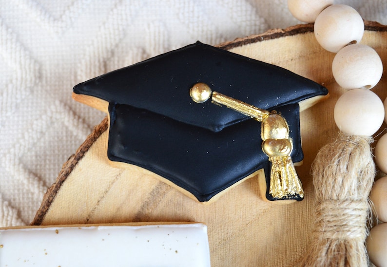 Pu&ograve; includere: Biscotto a forma di cappello di laurea nero decorato con glassa dorata e un fiocco. Il biscotto &egrave; su una fetta di legno con una ghirlanda di perline di legno.