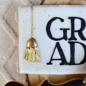Pu&ograve; includere: Biscotto quadrato bianco decorato con glassa nera e la parola "GRAD" e un fiocco dorato.