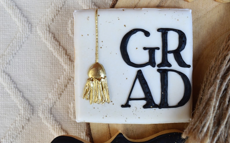Pu&ograve; includere: Biscotto quadrato bianco decorato con glassa nera e la parola "GRAD" e un fiocco dorato.