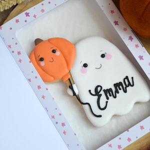 May include: A white ghost-shaped cookie with a smiling face and pink cheeks, holding an orange pumpkin-shaped balloon. The name Emma is written in black icing on the ghost. The cookie is presented inside a white box with a window and pink star pattern.