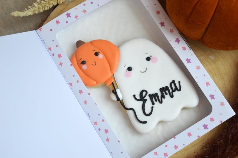 May include: A white ghost-shaped cookie with a smiling face and pink cheeks, holding an orange pumpkin-shaped balloon. The name Emma is written in black icing on the ghost. The cookie is presented inside a white box with a window and pink star pattern.