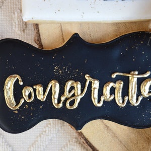 Pu&ograve; includere: Biscotto nero e dorato decorato con la parola "Congrats" in glassa dorata. Il biscotto ha la forma di un cartello.