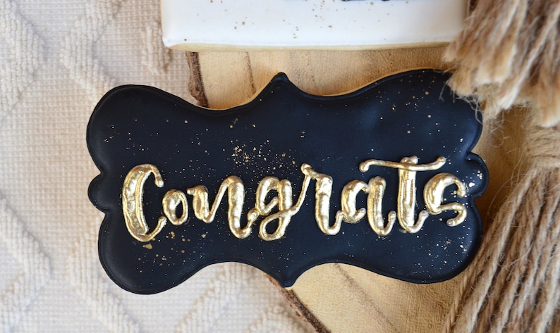 Pu&ograve; includere: Biscotto nero e dorato decorato con la parola "Congrats" in glassa dorata. Il biscotto ha la forma di un cartello.