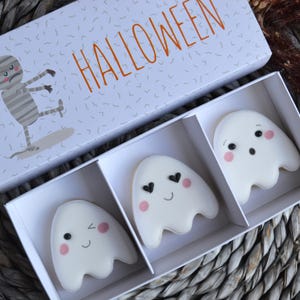 Pode incluir: Uma caixa retangular branca com o texto laranja HALLOWEEN e uma ilustração de múmia, contendo três biscoitos brancos em forma de fantasma. Cada biscoito apresenta um glacê liso, olhos pretos e bochechas rosadas, com expressões faciais variadas.